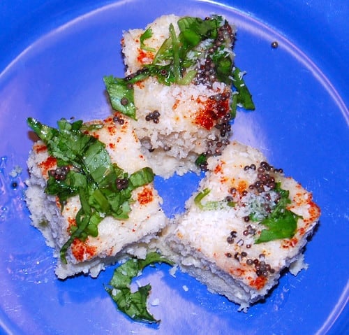 Photo of three pieces of brown rice dhokla with cayenne, mustard seeds and cilantro on a blue plate.