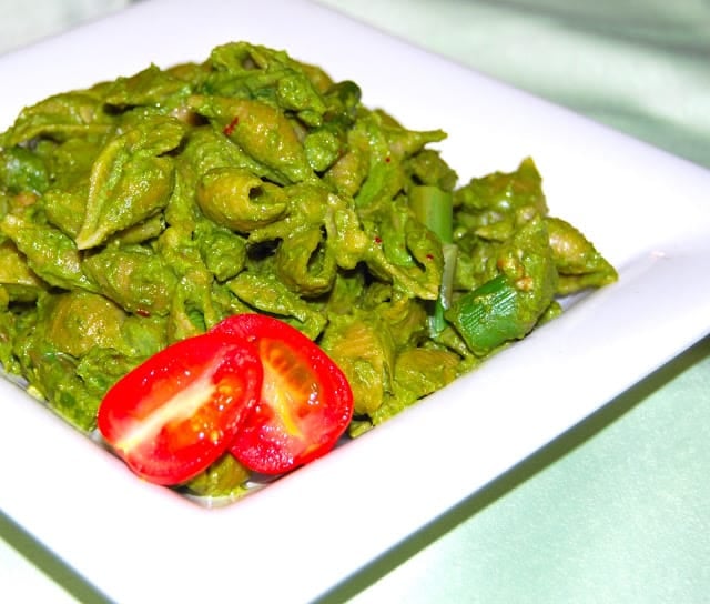 Photo of a plate of conchiglie or shell pasta with a green, fresh, vegan kale pesto and a slice of tomato.
