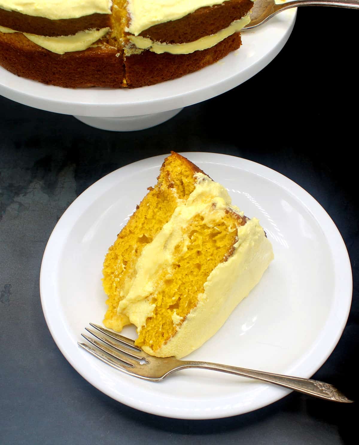 Photo of a vegan mango cake slice on a white plate with the whole cake in the background on a cake stand.