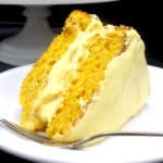 A luscious slice of vegan mango cake with smooth, creamy mango buttercream frosting on a white plate with a fork.