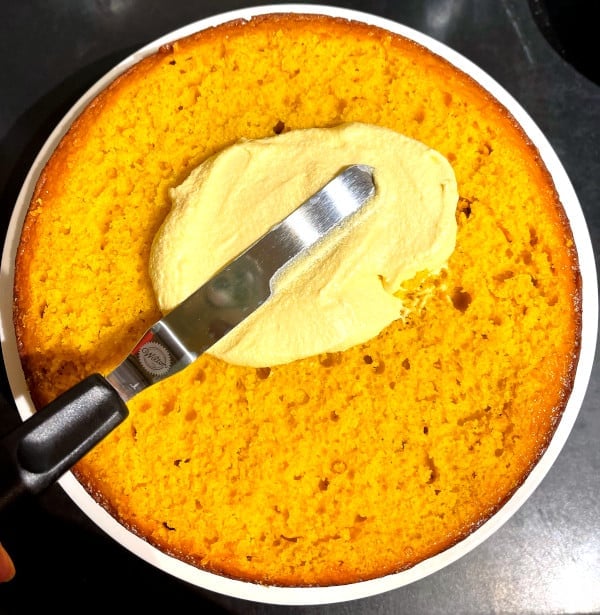 Icing being spread over mango cake with offset spatula.