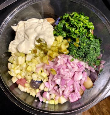 Ingredients for vegan potato salad in glass bowl, including vegan mayo, shallots, celery, parsley, dill, pickles and mustard.
