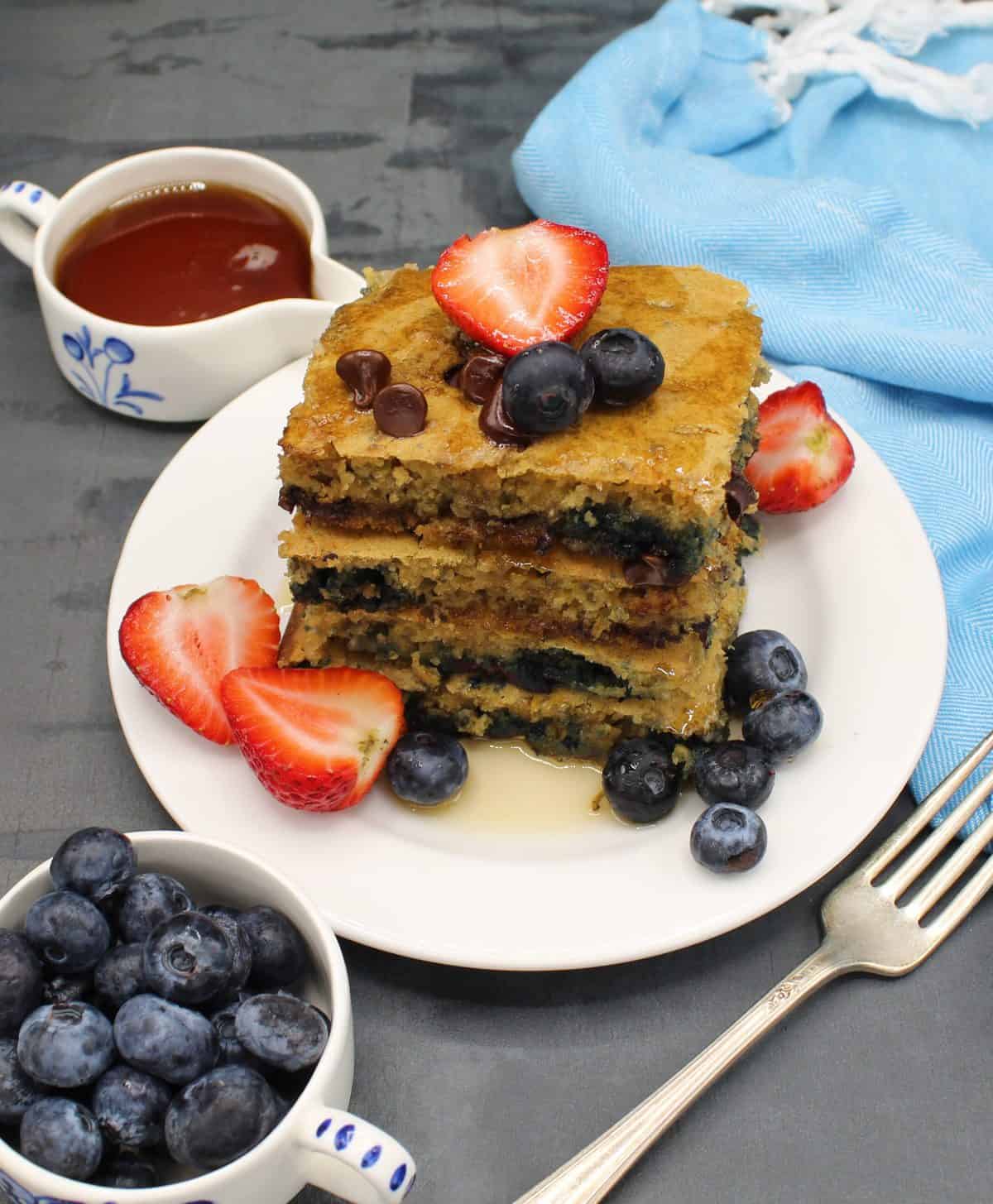 Four vegan sheet pan pancake squares stacked on a white plate with maple syrup and berries and a fork and blue napkin on the side.