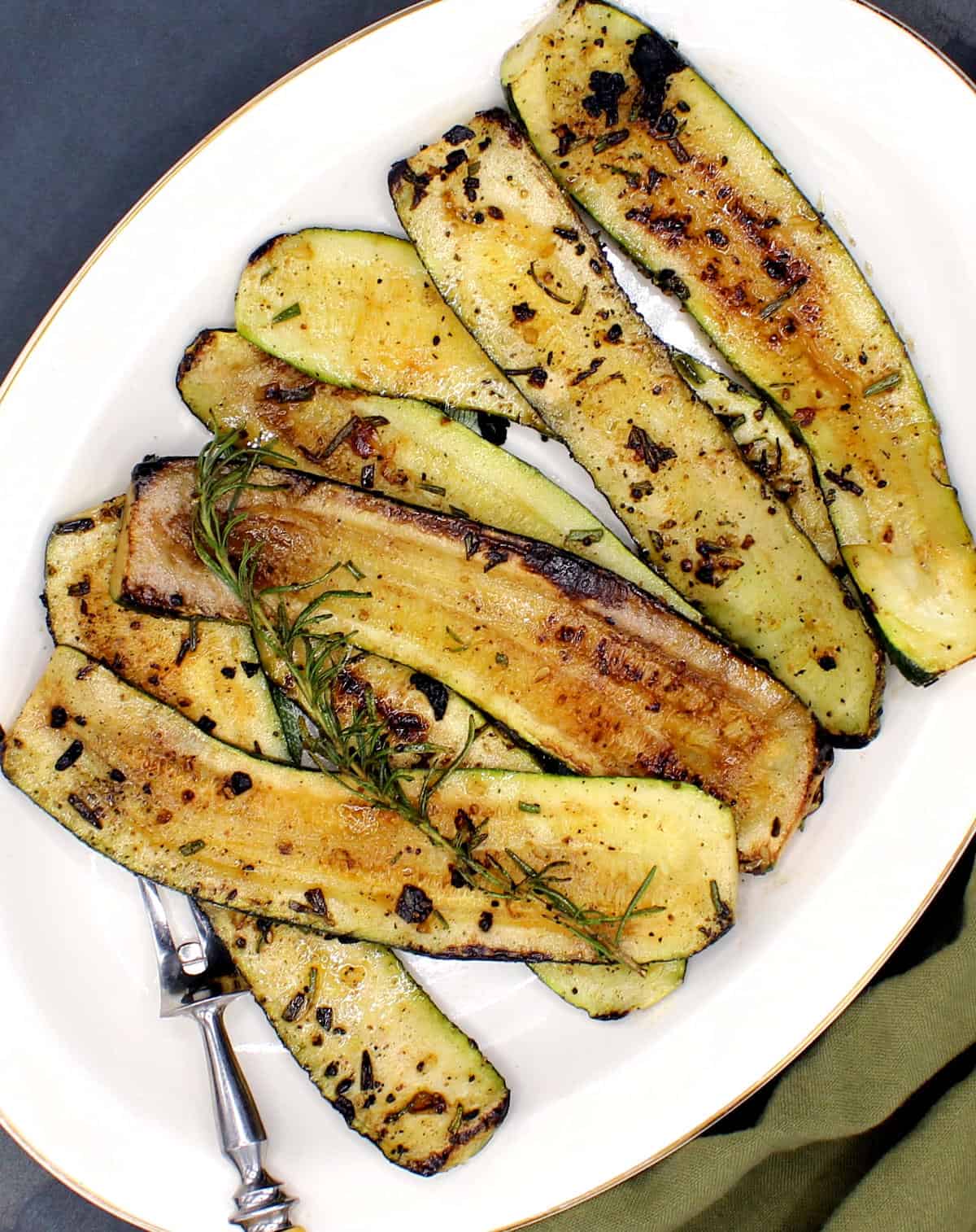 Overhead photo of slices of pan roasted, golden-brown zucchini in a white platter with a rosemary sprig.