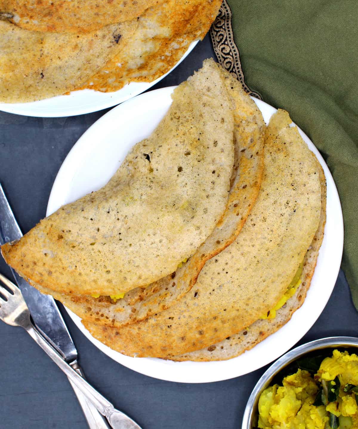Overhead photo of two sorghum dosas on a white plate with more dosas and potato sabzi around them.