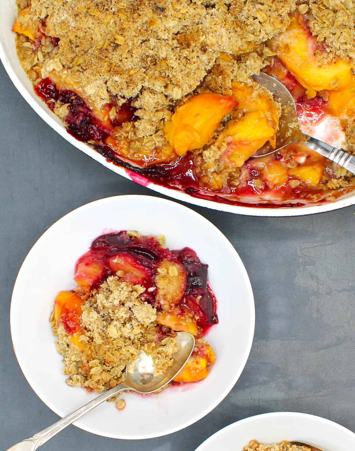 Vegan Peach and Plum Crisp in a white bowl with a spoon and the baking dish with the crisp and pieces of peach.