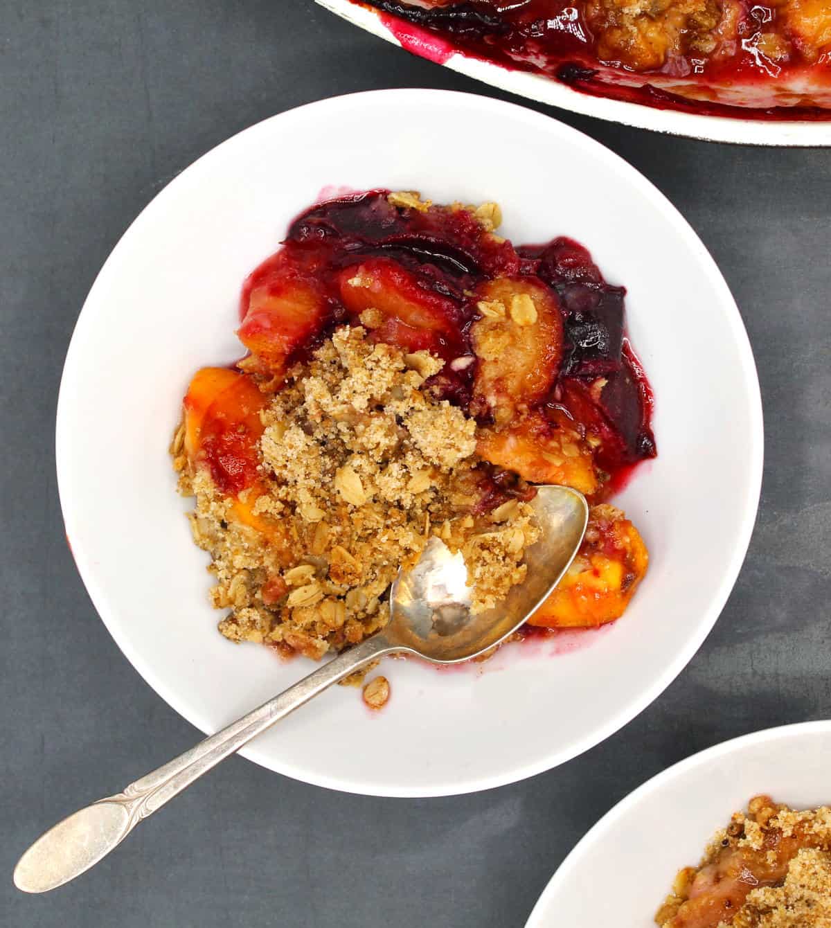 Closeup of vegan peach and plum crisp in a white bowl with spoon.