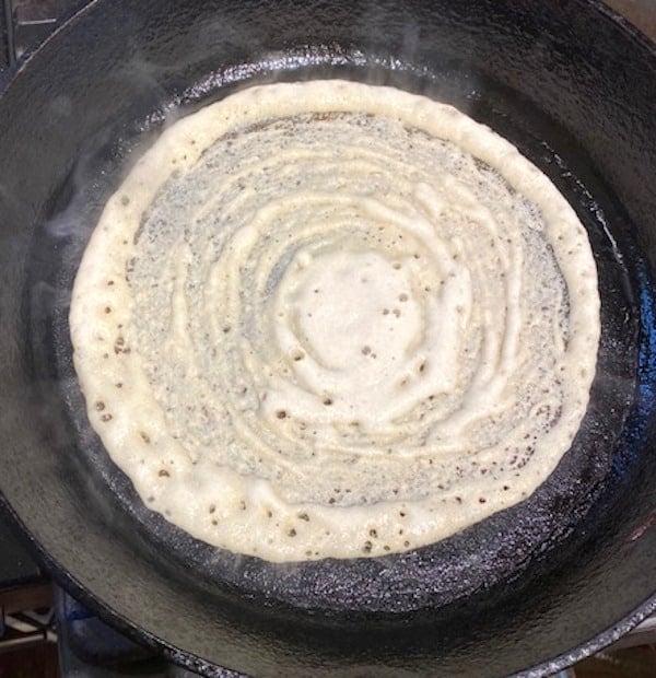 Quinoa dosa being cooked on a cast iron griddle.
