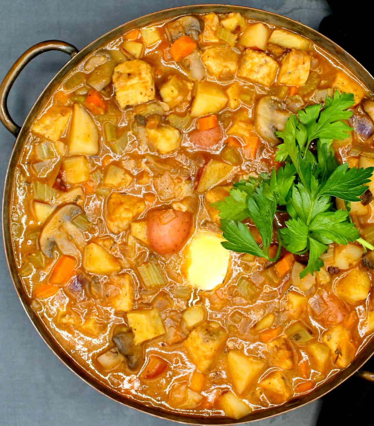 Overhead shot of a copper serving dish with vegan veggie-packed stew with tempeh, carrots, potatoes, onions, celery and carrots and a garnish of parsley.