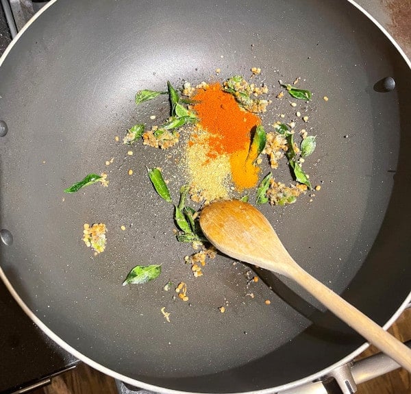 Wok with hot oil and spices added to it with wooden ladle.