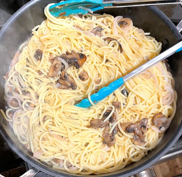 Spaghetti added to sauce with mushrooms and shallots in saute pan.