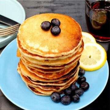 A tall stack of vegan lemon pancakes with lemon slices and blueberries on a blue plate.