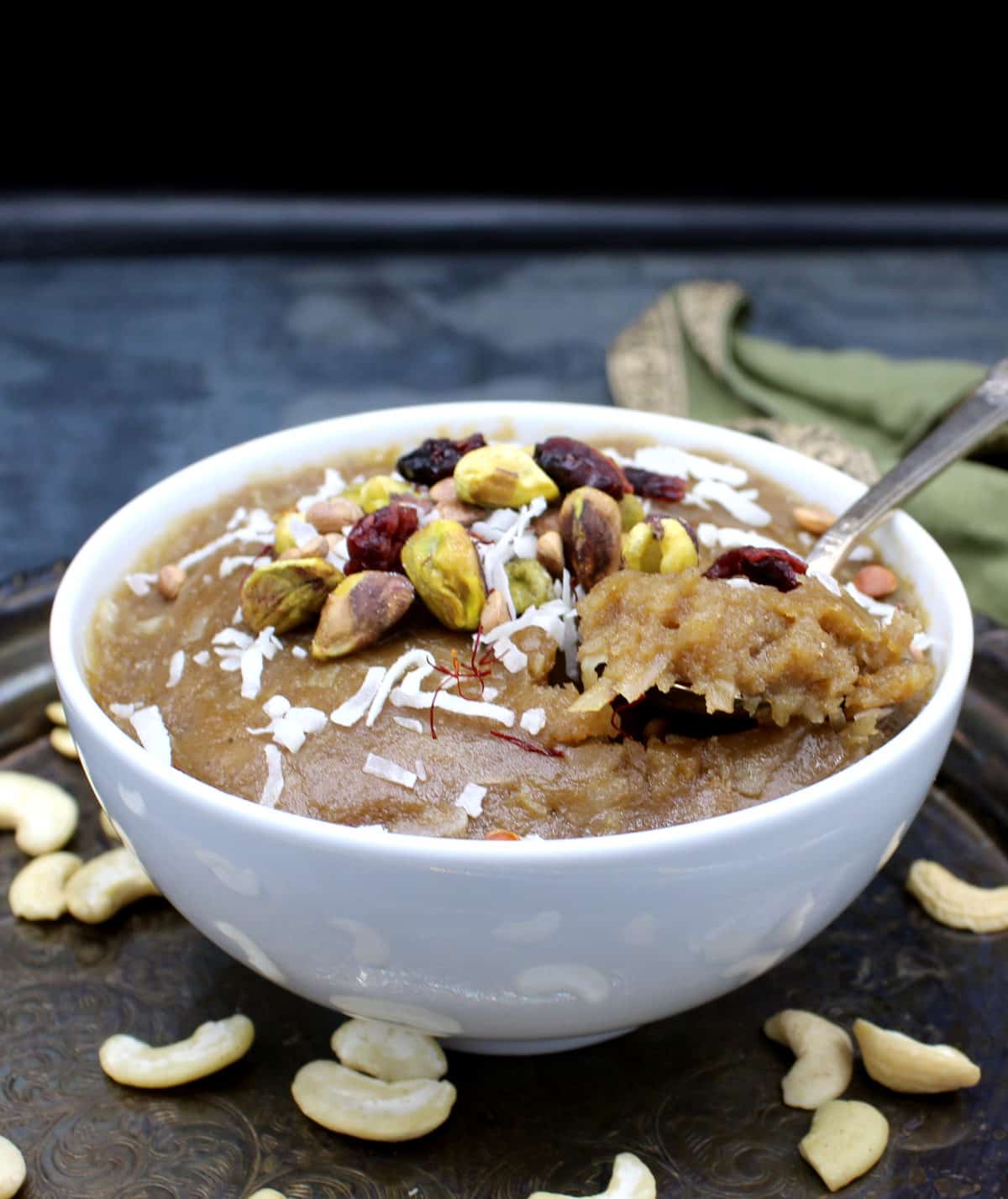 Front photo of a bowl of vegan cashew halwa garnished with nuts, dry fruits and coconut flakes with a green napkin.