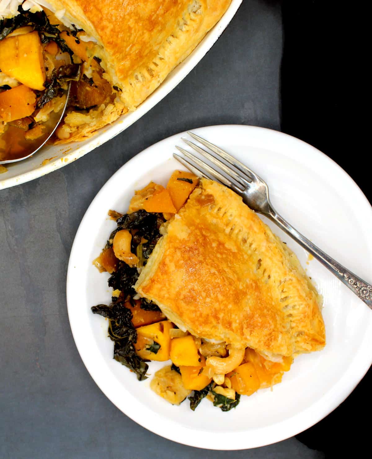 Overhead shot of a slice of vegan butternut squash pie with kale and cashews on a white plate with fork and partial view of full pie in background.