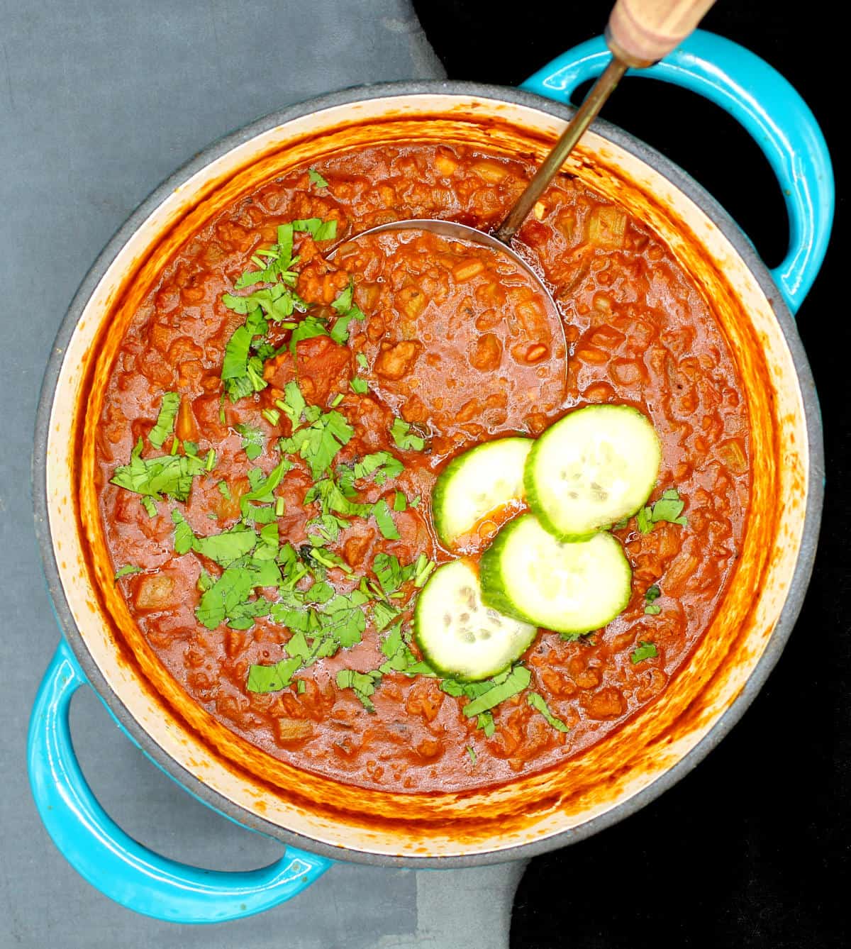 Overhead shot of vegan keto low carb chili with parsley and cucumber slices in a teal dutch oven with ladle.