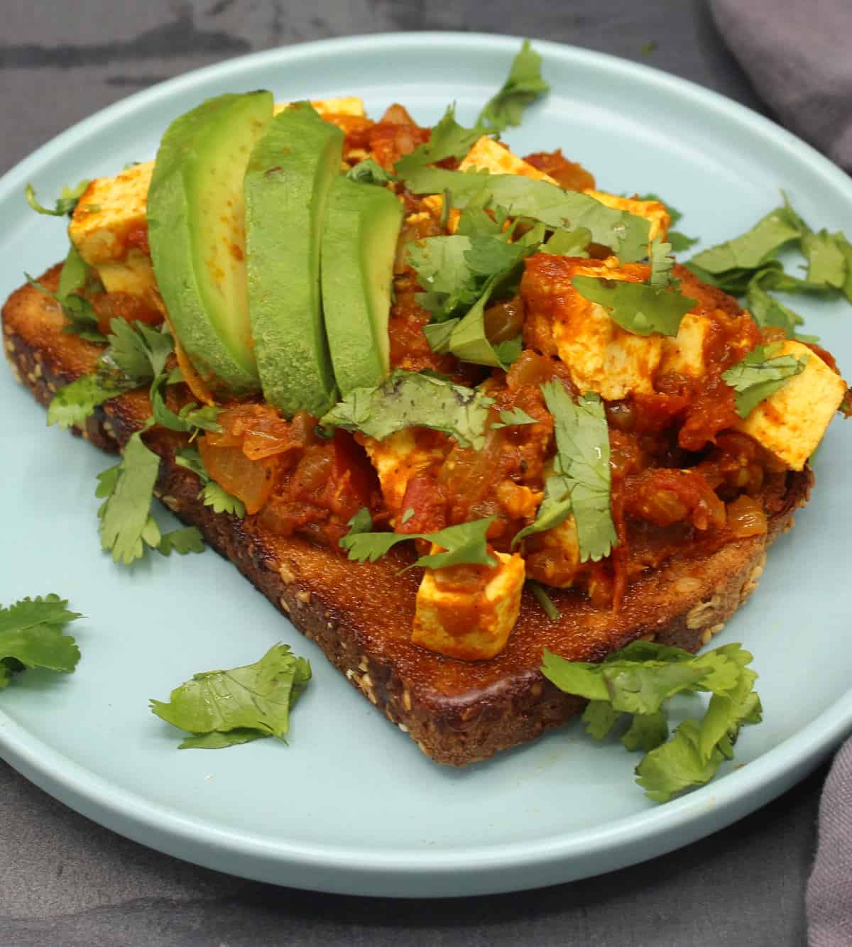 Tomatoes with Tofu served over toast with slices of avocado and a cilantro garnish.