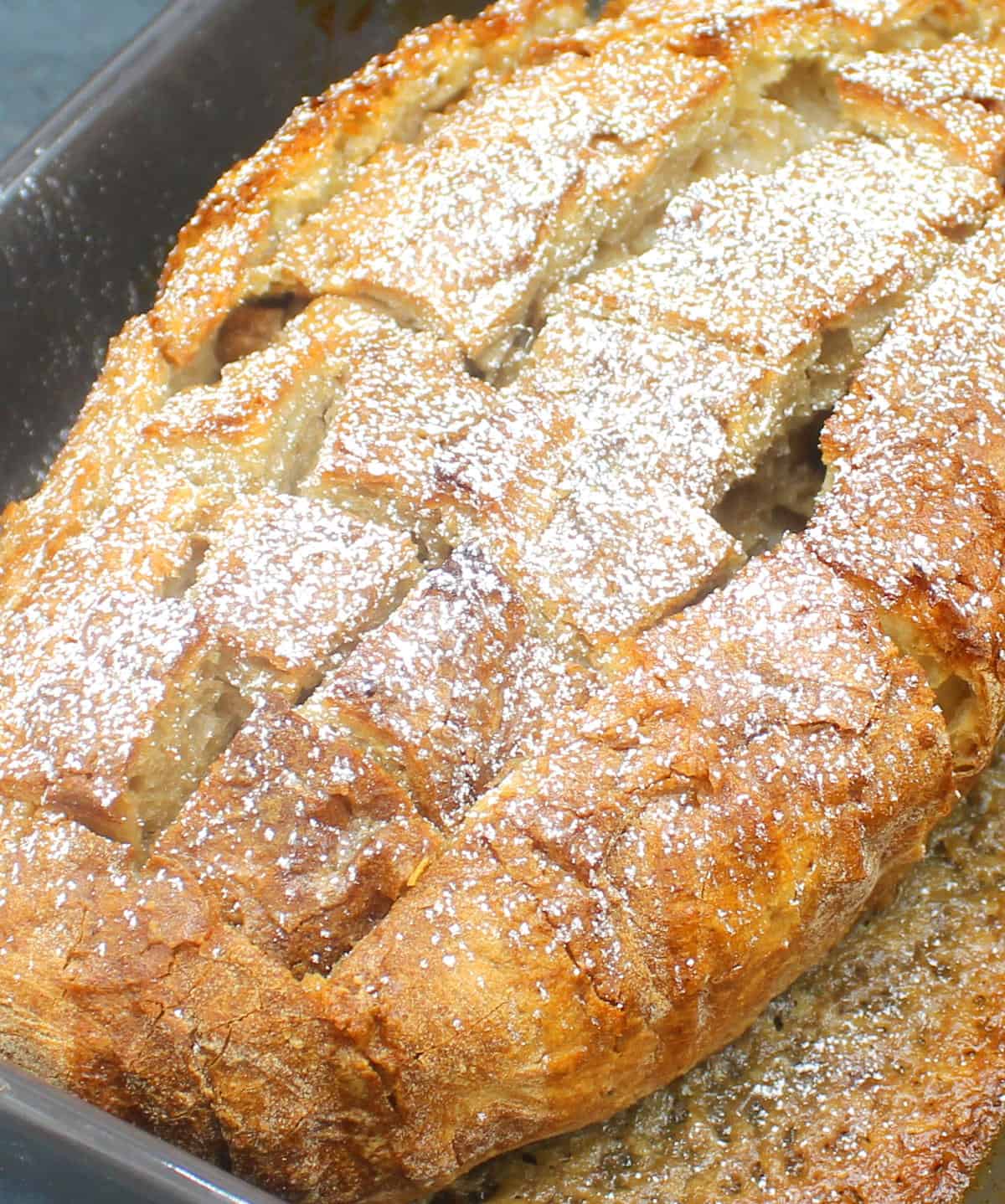Front closeup of a loaf of vegan eggnog french toast with square cut into it in baking dish and powdered sugar dusted over.