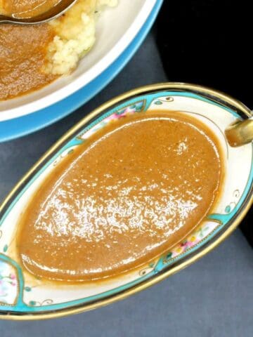 Vegan gravy in sauce boat with partial view of mashed potatoes and gravy in background.