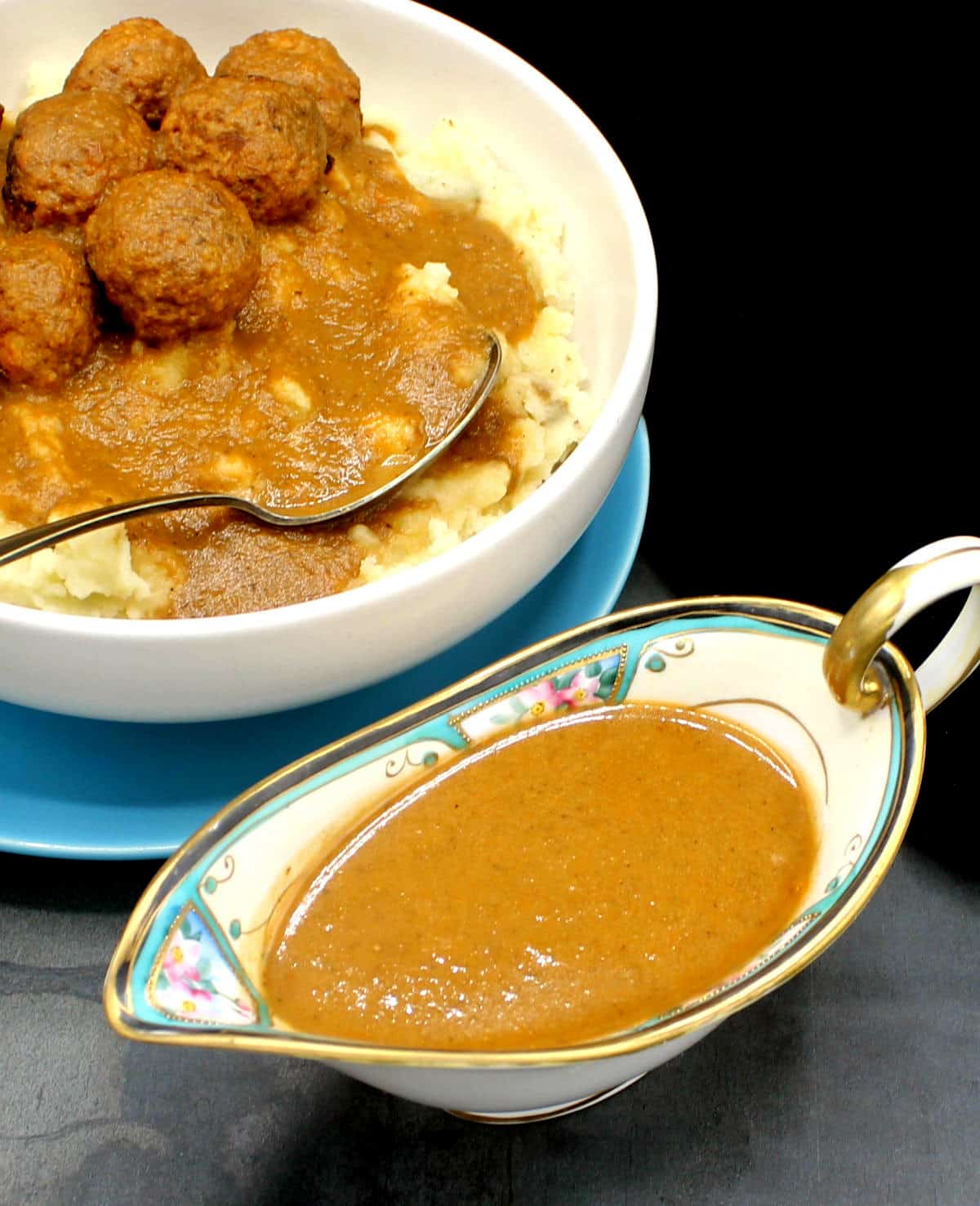 Front photo of vegan gravy in an ornate sauce boat with a bowl of vegan meatballs and mashed potatoes with gravy in the back.