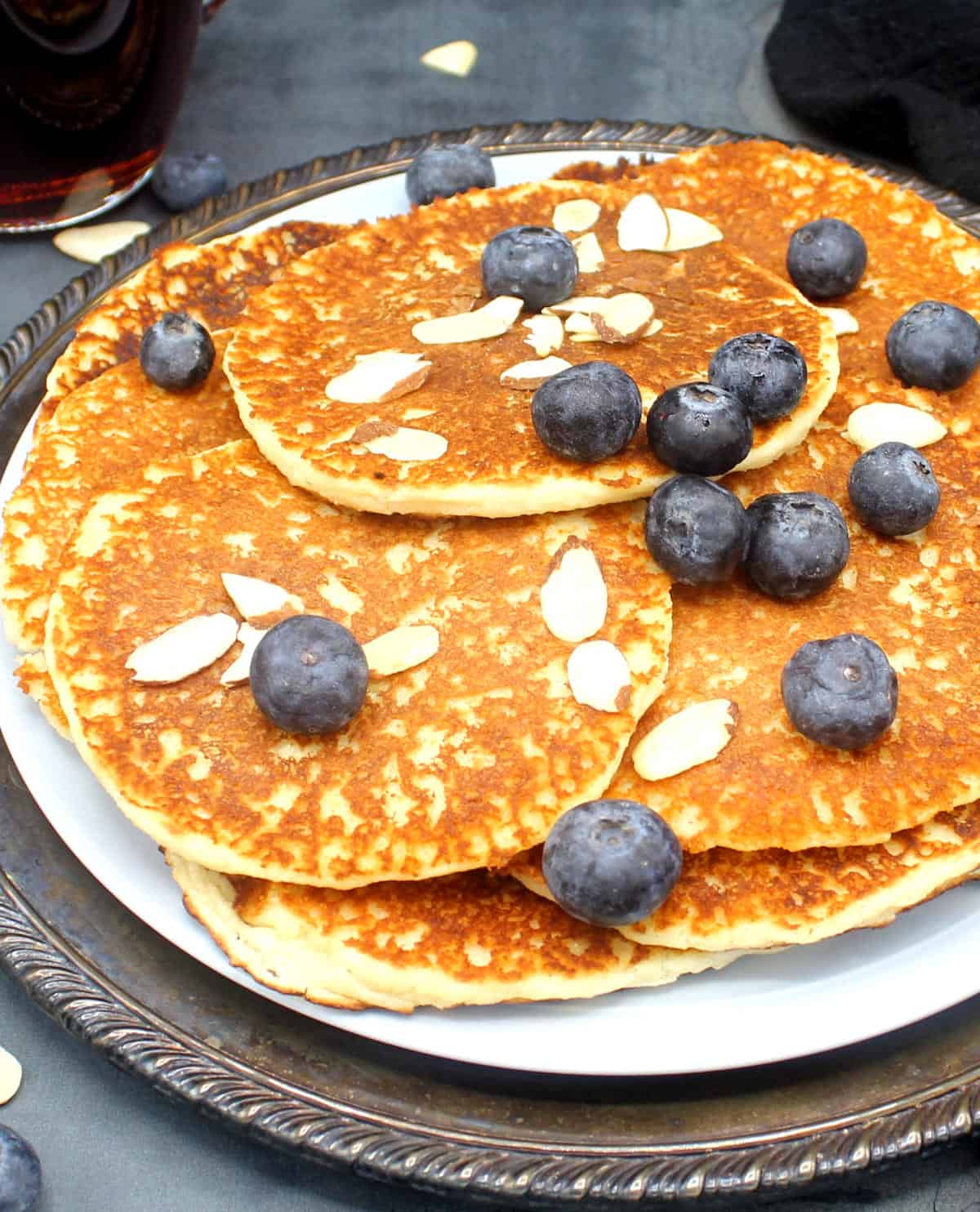 Front partial shot of a pile of vegan low carb pancakes in a blue plate with blueberries and sliced almonds with maple syrup in background.