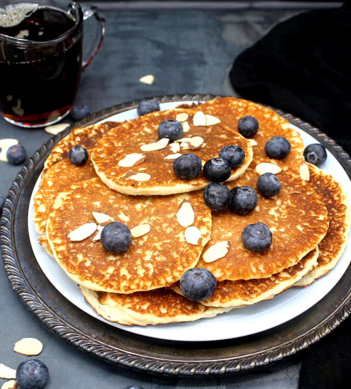 Front photo of vegan keto low-carb pancakes in a plate with sliced almonds and blueberries and maple syrup in background.