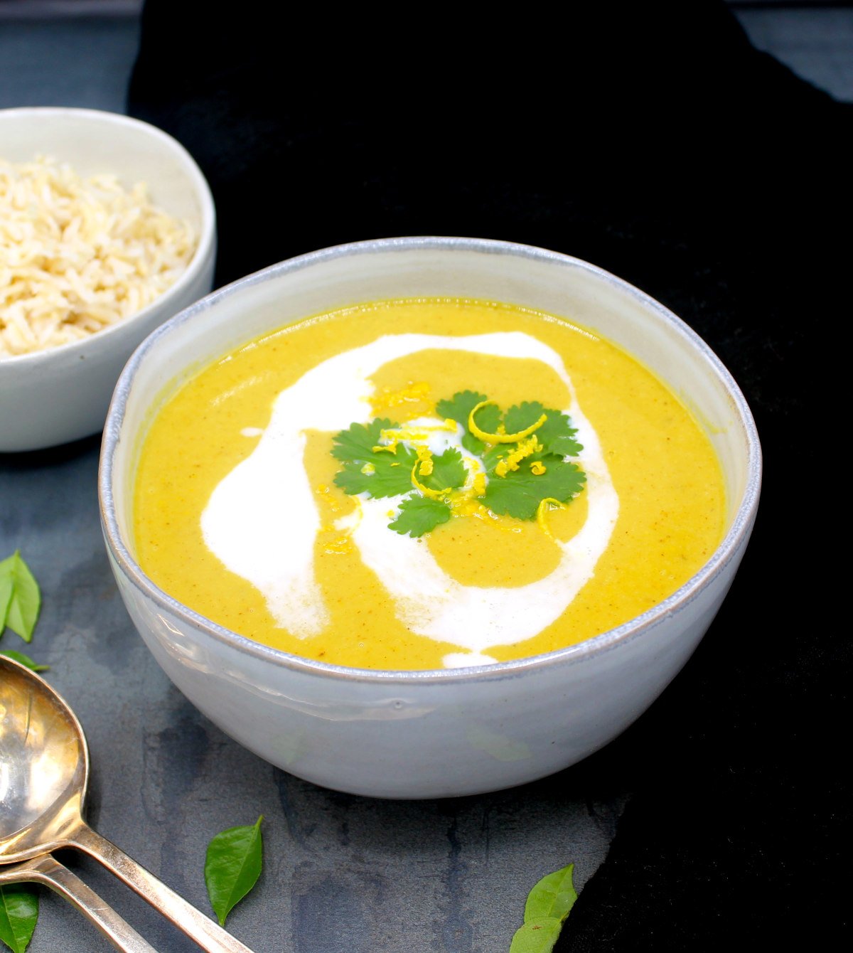 Front photo of a gray bowl with creamy mulligatawny soup with coconut cream, cilantro and lemon zest. On the side is a bowl of brown rice and soup spoons with curry leaves scattered around.
