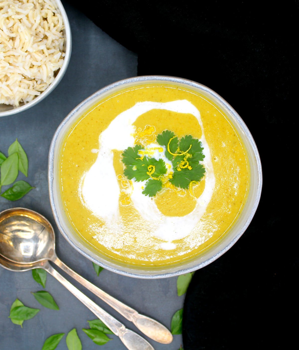 Overhead shot of a bowl of mulligatawny soup with coconut milk, cilantro, lemon zest and a bowl of rice on the side. Also in the picture are soup spoons and curry leaves scattered around.