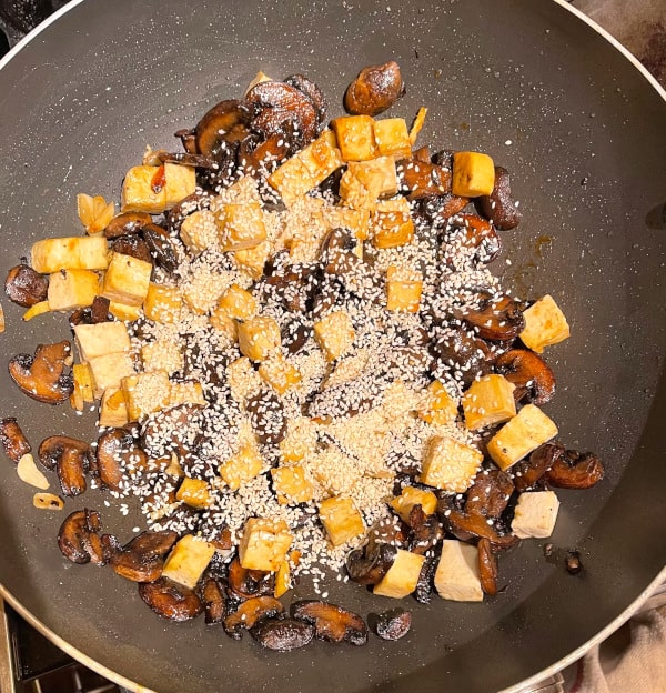 Sesame seeds added to tofu and mushrooms in wok.