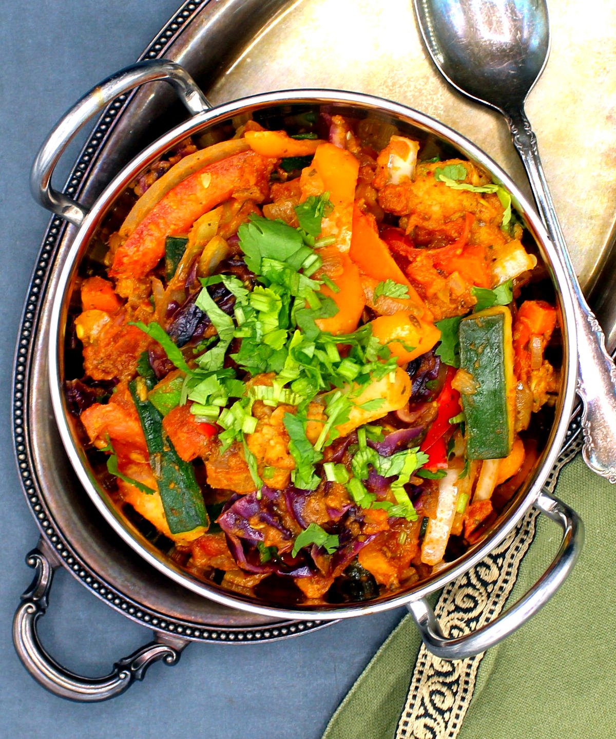 Overhead shot of mixed vegetable jalfrezi in a kadhai over a silver tray with a spoon and green napkin.