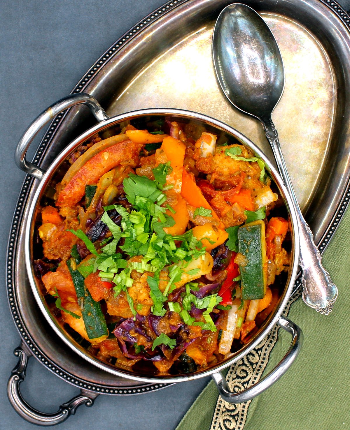 Overhead shot of colorful vegetable jalfrezi in a kadhai with a spoon and green napkin next to it.