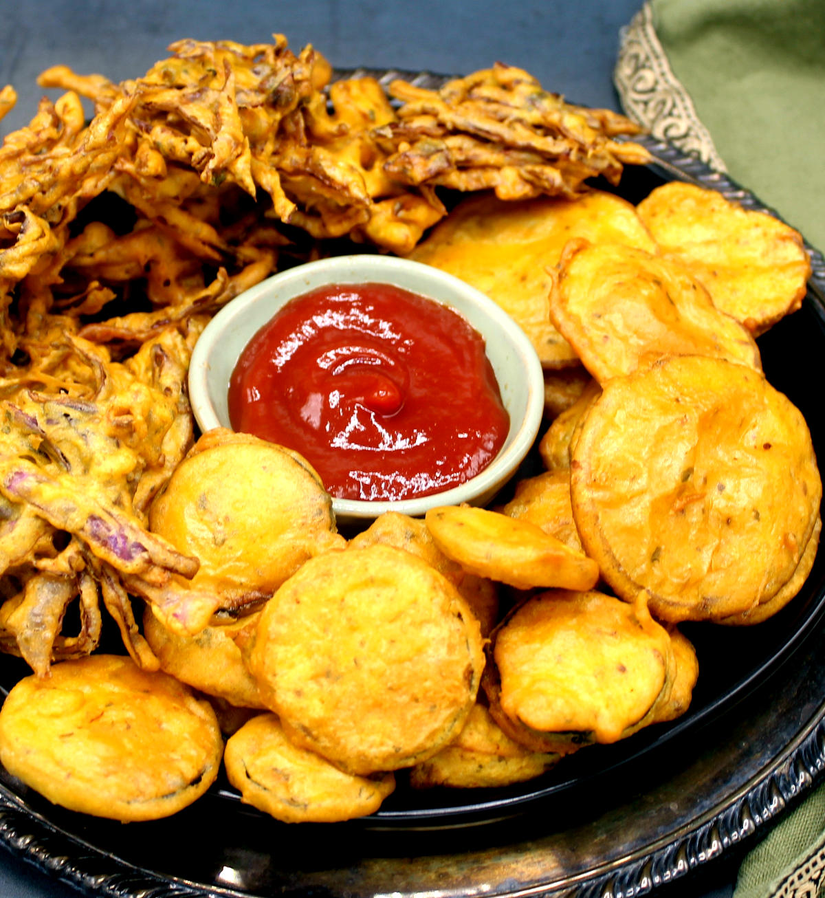 Front partial closeup photo of a plate packed with vegetable pakora or bhajias or pakodas with maggi masala sauce and a green napkin.