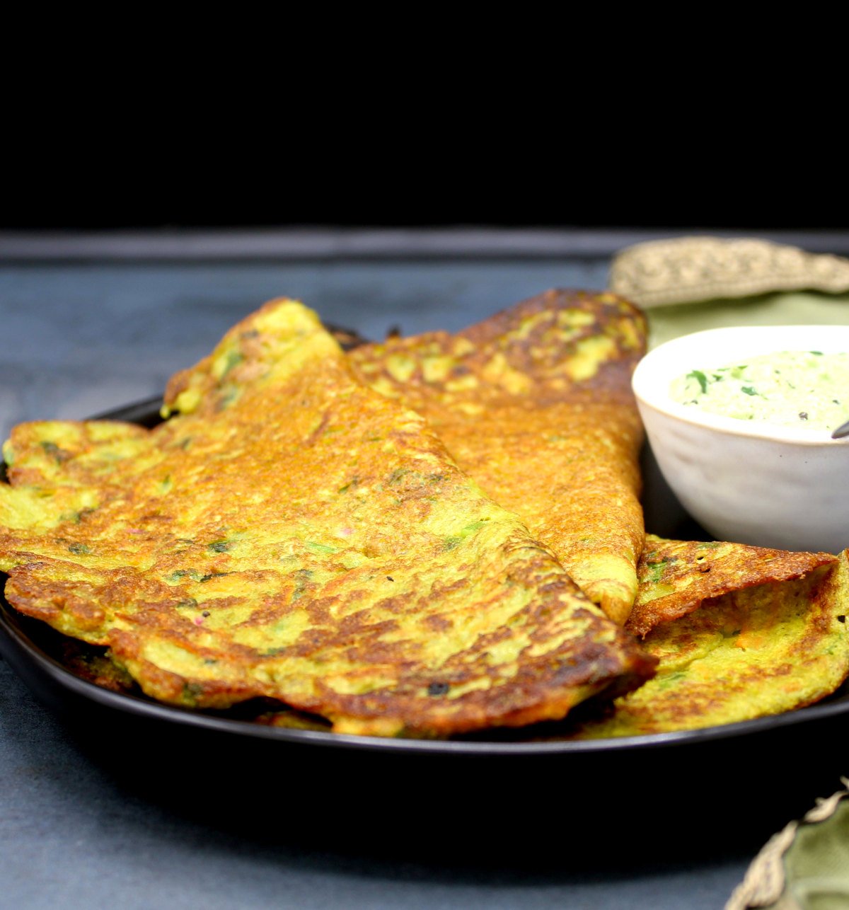 Front partial shot of golden brown moong cheelas in a black plate with chutney.