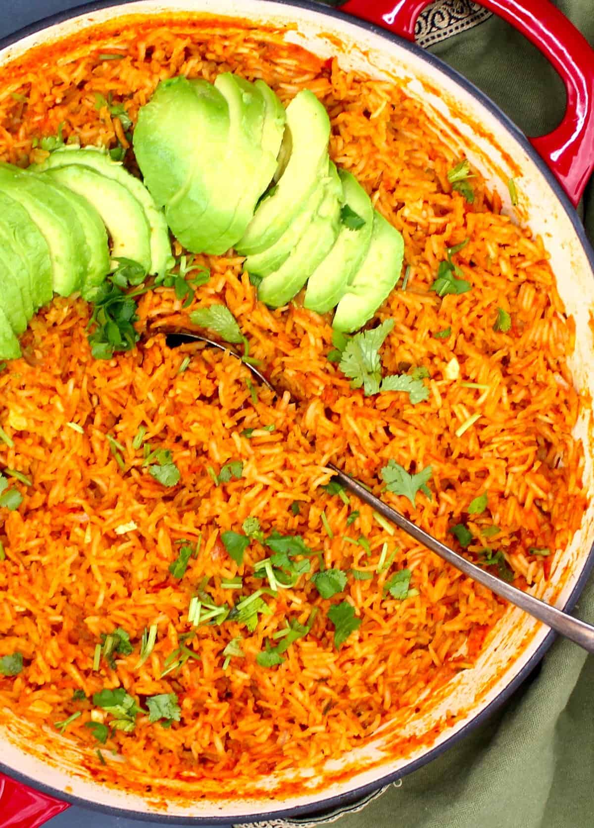 Partial overhead photo of Mexican tomato rice in skillet with a spoon, cilantro and avocados.