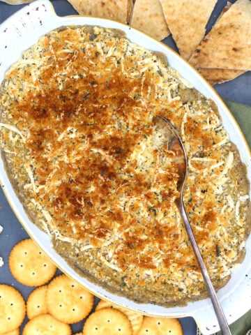 Hot vegan spinach dip in baking pan with spoon and with crackers strewn around.