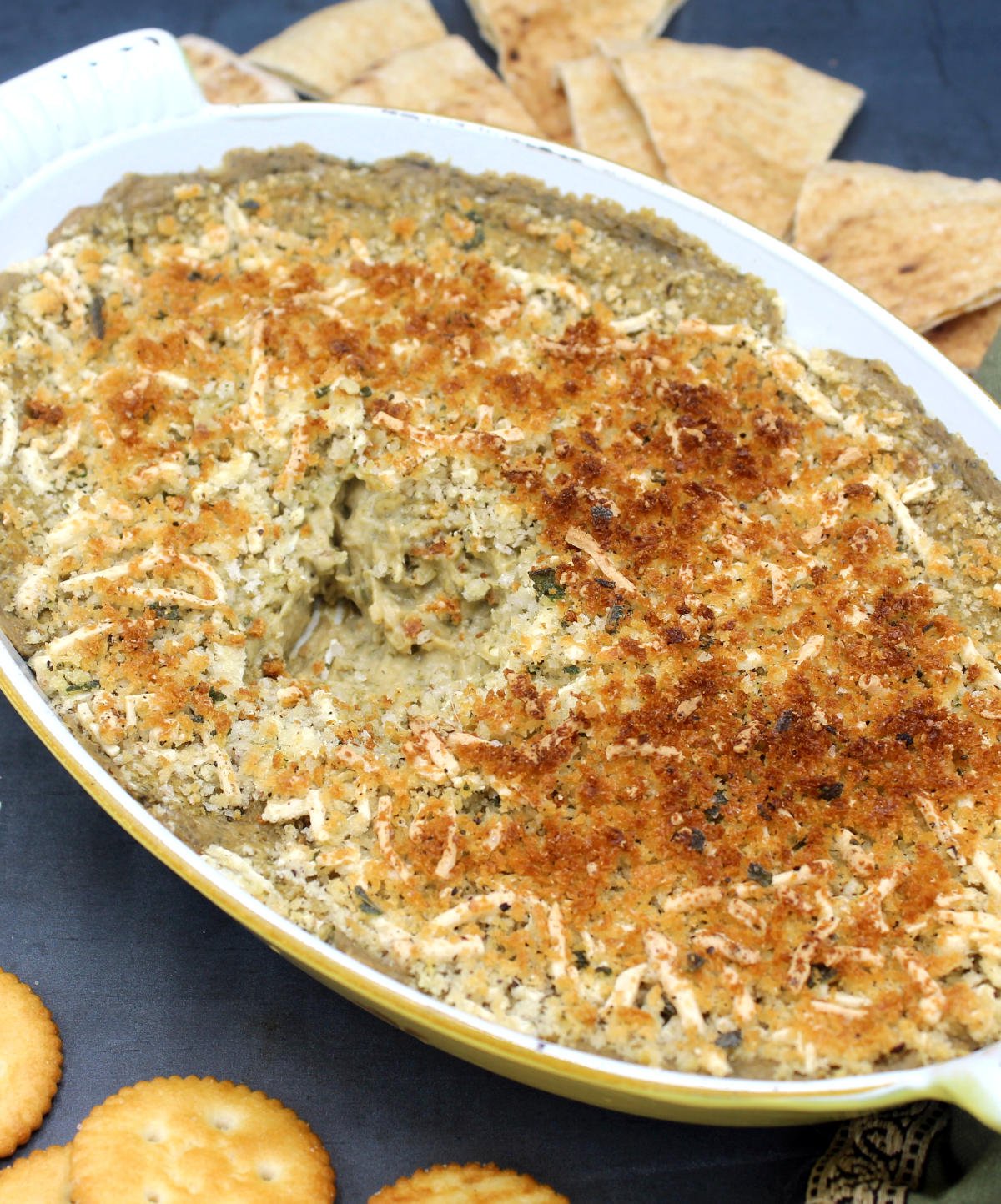 Front partial closeup of vegan spinach dip with caramelized onions in a baking dish surrounded by crackers and pita bread.
