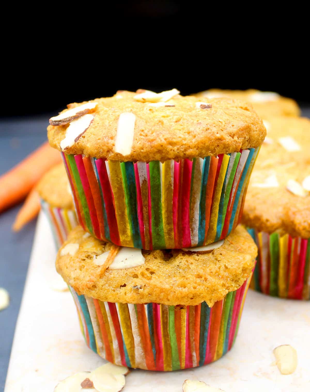 Two carrot muffins with colorful liners and studded with almonds stacked on top of each other with more muffins and carrots in background.