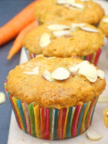 Image of a vegan carrot muffin on marble chopping board with more muffins in background, and carrots and almonds scattered around.