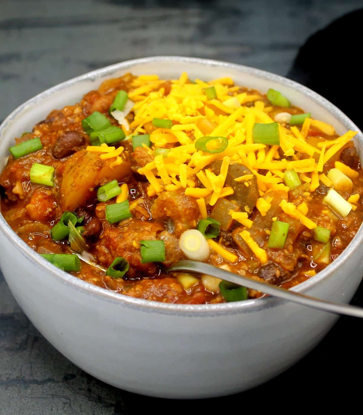 Front photo of a gray ceramic bowl with vegan Irish chili with a garnish of scallons and vegan cheddar cheese shreds.