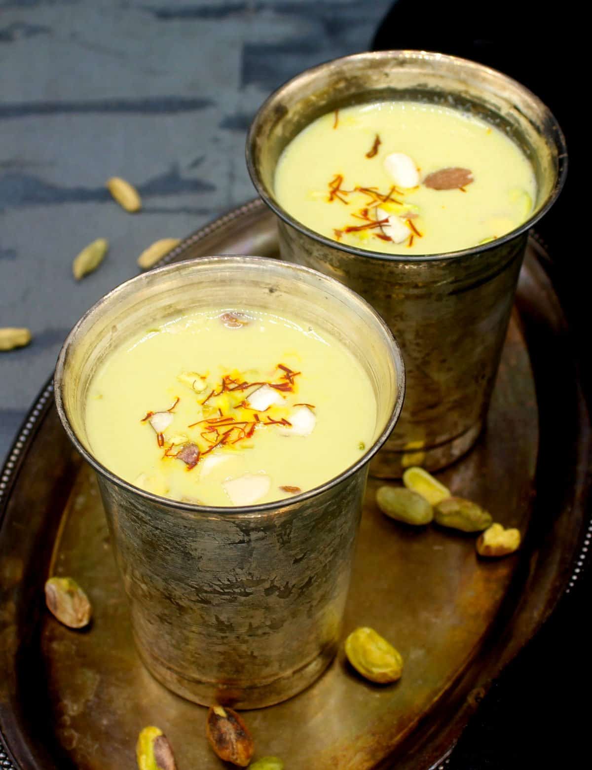Front shot of two silver glasses filled to the brim with vegan thandai with saffron and nuts garnish and nuts and cardamom pods scattered around.