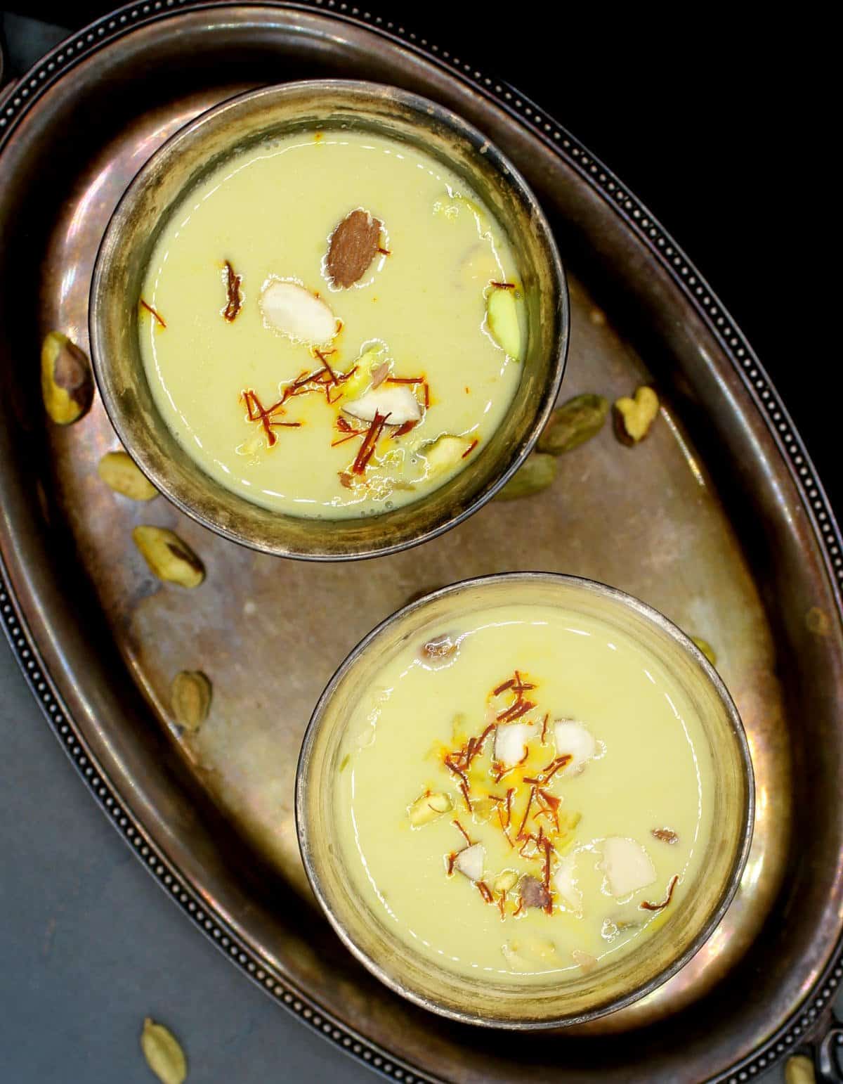 Overhead shot of two silver glasses of thandai with oat milk, saffron strands, almonds and pistachios.