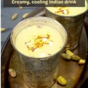 Two silver glasses with vegan thandai on silver tray with nuts scattered around.