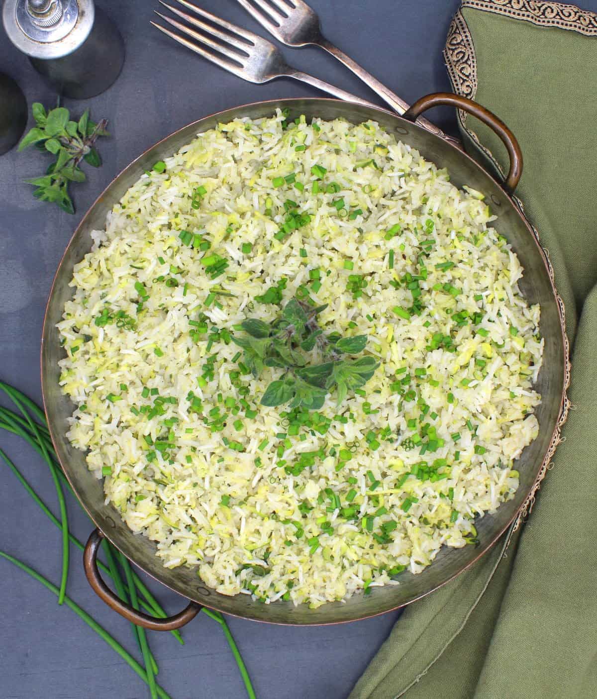 Overhead view of garlicky zucchini rice in a copper pan with chives and oregano garnish with more chives, oregano, forks and a salt and pepper shaker in background.