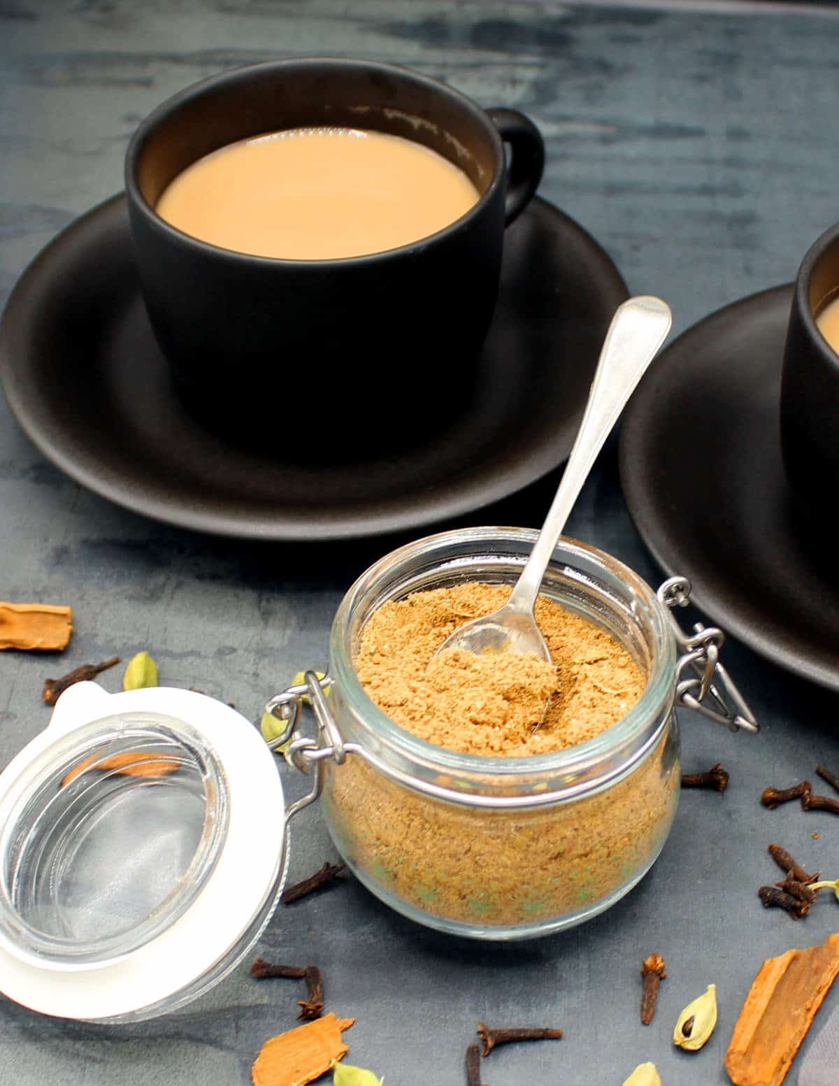 A glass jar with chai masala and spoon with cups of masala tea in background and spices lying around.