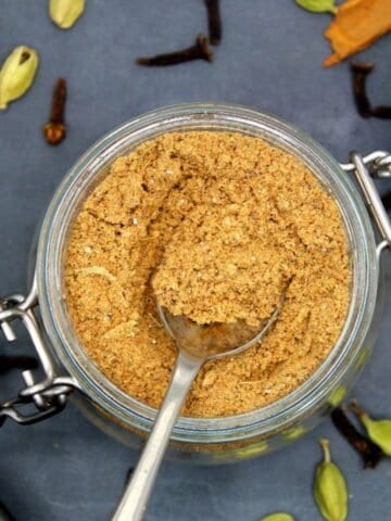 Chai masala with spoon in glass jar with spices strewn around.