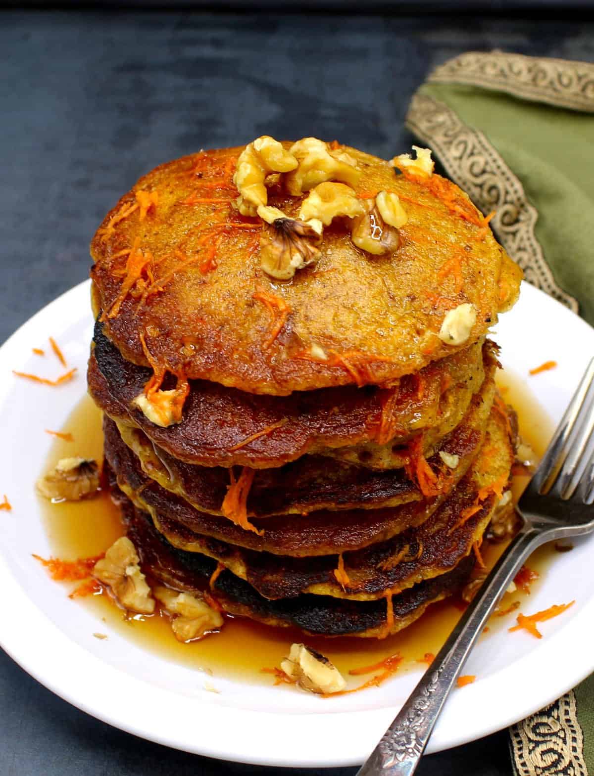 A stack of vegan carrot cake pancakes on a white plate with shredded carrots and chopped walnuts. Next to the pancakes is a silver decorative fork and a green napkin is next to plate.