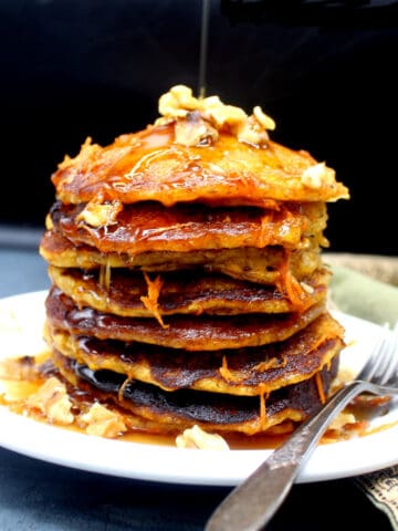 Vegan carrot pancakes stacked on a plate with nuts and maple syrup.