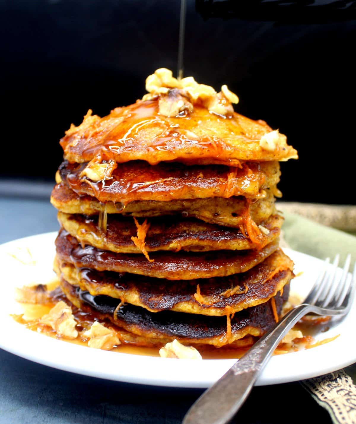 A stack of vegan pancakes on a white plate with walnuts and shredded carrots with a fork. Maple syrup is being drizzled over.