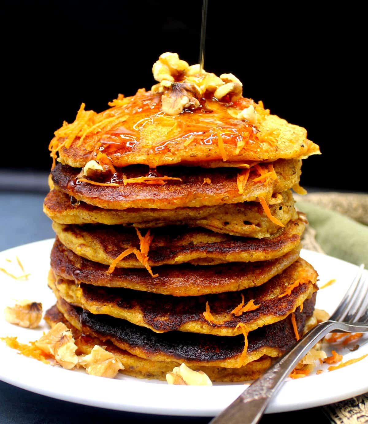 A stack of vegan carrot cake pancakes with walnuts and shredded carrots in white plate with walnuts and maple syrup being drizzled over it.