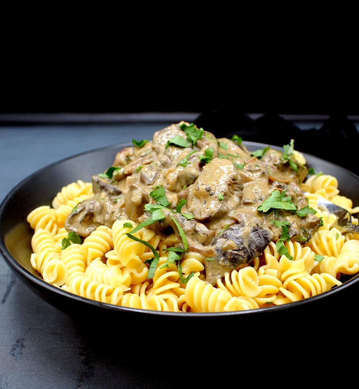Front shot of vegan mushroom stroganoff over corkscrew pasta in black bowl.