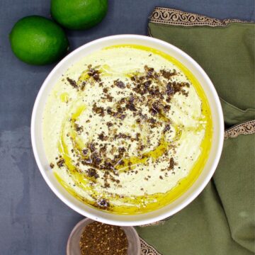Houmous edamame crémeux dans un bol blanc avec une serviette verte, deux citrons verts et du za'atar dans un petit bol qui l'entoure.